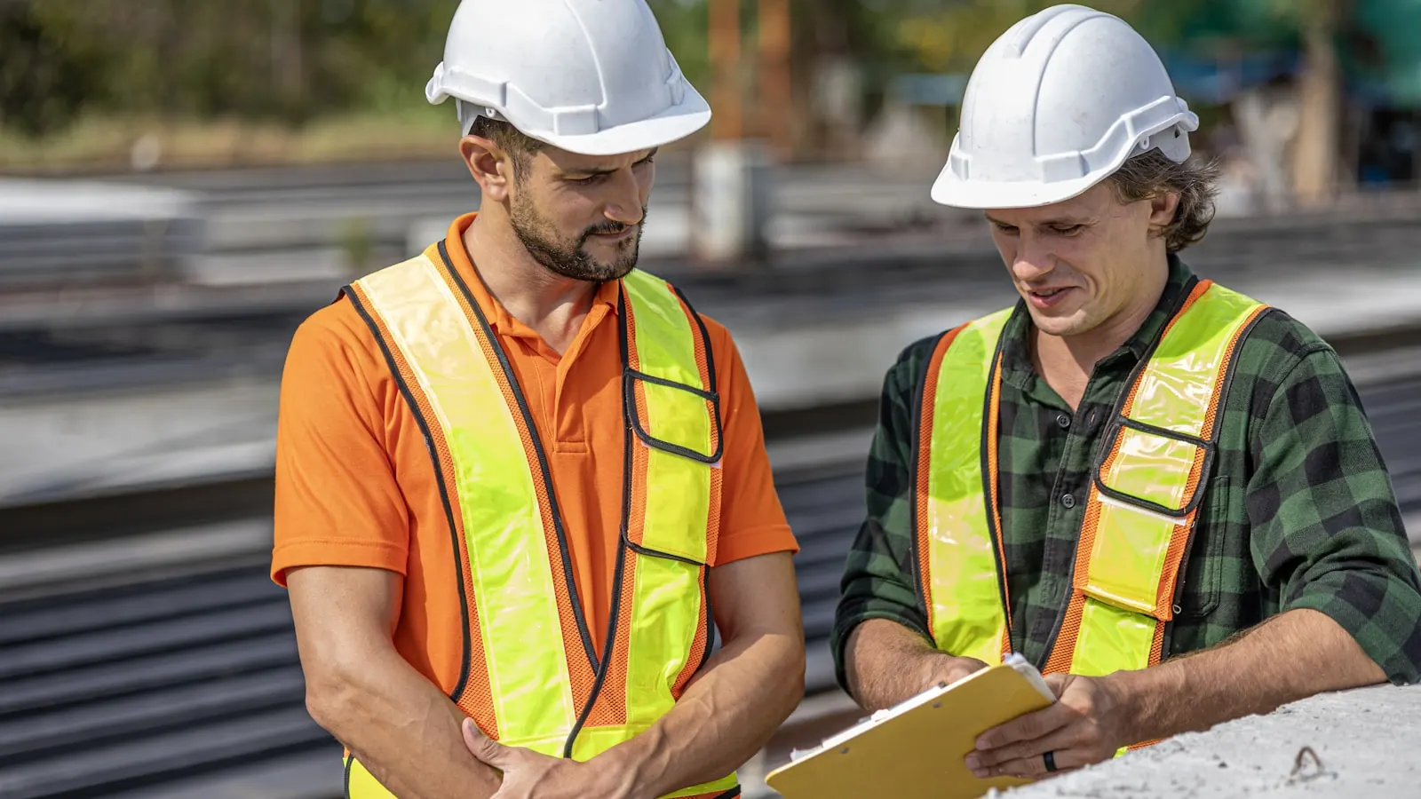 Two workers in hard hats and hi-vis vests going through paperwork on a clipboard together on a construction site